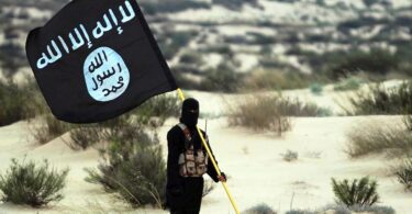 A masked man in the desert in either Iraq or Syria while holding the flag of the Islamic State in Iraq and Syria (ISIS)